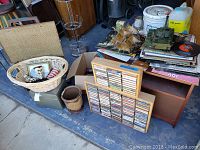 Wide shot showing wooden table with records, cassette tapes, wicker basket with small items, toy tank, and model ship on and around the table.