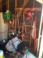 Photograph showing the red leaf blower, two trimmers, Scotts spreader seeder mounted on two wheels with green hopper, and other gardening equipment stored in a shed setup.