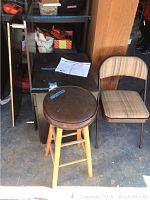 Unassembled pieces of a desk and filing cabinet including instructions, with a brown round stool with wooden legs, and a folding chair with a striped fabric seat and backrest, placed on a concrete floor.
