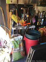 Photo of white broom rack with multiple brooms, mops, rakes, crowbars, bungee cords, dust pans and other yard and garden tools placed on and around a white table along with two large red and black garbage cans and bags of planting soil.