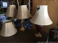 Three table lamps on wooden surface: two have matching brass bases and fabric shades with embroidery, the third has a ceramic base with a brass bottom and a cream shade with gold trim. Extra shade included.