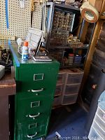 Green metal file cabinet with three drawers, surrounded by plastic storage drawers and a small hardware compartment organizer on a pegboard wall.