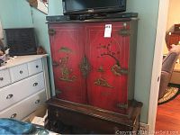 Front view of red antique Asian inspired cabinet with gold nature designs and metal hardware, standing on dark wooden base, showing label and TV on top.