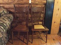 Pair of vintage wooden chairs side by side against wooden wall and floor, showing carved decorative backs and vertical slats.