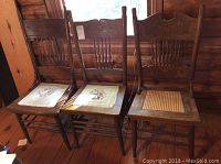 Three vintage wooden chairs placed side by side on wooden floor, showing two needlepoint seats and one cane seat.
