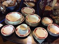 Photo of multiple stacked plates, bowls, saucers, and a coffee cup with decorative red floral rim design on a round table.