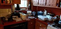 View of countertop showing multiple kitchen appliances including toasters, waffle irons, sandwich grill, electric knife, and small kitchen tools scattered on counter in a wooden kitchen setting.