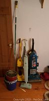 Four cleaning appliances lined up against wall: Blue vintage Eureka The Boss upright vacuum, yellow Eureka The Boss Super Lite upright vacuum, white Eureka The Boss Smart Vac upright vacuum, Bissell steam mop handle with mop head attachment. Several buckets stacked nearby.