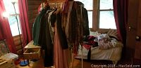 Image showing a wooden room interior with several coats, dresses, and scarves hanging on a clothes rack alongside a bed with cushions and folded clothes