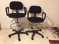 Two black captain's style bar chairs with armrests, adjustable height with footrest and caster wheels, placed on carpeted floor against a white brick wall.