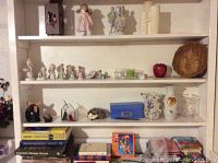 Shelf with Precious Moments and Growing Up figurines, several decorative vases, candle holders, a red apple decor piece, and a woven basket.