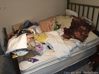 View of assorted linens, pillows, and throws piled on twin bed frame, showing variety of folded and loosely stacked textile items.