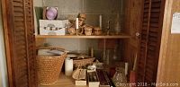 Open wooden cabinet showing oil lamp chimneys, candles, small boxes, and baskets on shelves