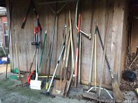 Photo showing a row of assorted yard tools leaning against a wooden wall including shovels, rakes, brooms, and digging tools.