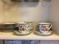 Photo of 4 tea cups with matching saucers and stacked dessert plates inside cabinet shelf.