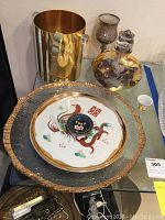 Photo of the large gold-tone metal ice bucket, ornate perfume bottle with floral decoration, and decorative serving platter with dragon design and gold trim on a glass surface.