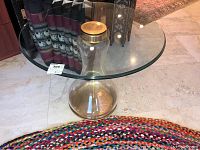Glass accent table with a round clear glass top and a fillable glass base shaped like a bottle with a gold-colored cap, shown on tile flooring next to colorful braided rug and other furnishings.