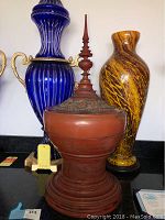 Front view of the wooden Tibetan rice server showing the tall spire and ornate lid with floral carving, flanked by decorative blue and yellow vases not included.