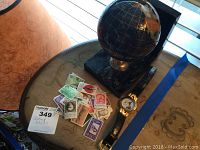 Display of the black globe paperweight on a square black base, assorted postage stamps spread on the table, and the Christopher Columbus watch with leather strap placed beside them.