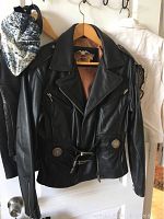 Front view of black Harley Davidson leather jacket hanging on wooden hanger. Features zipper pockets, collar, and belt with buckle.