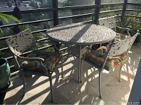 Wide shot of cast iron patio table and four matching chairs with cushions in outdoor setting with sun casting shadows.