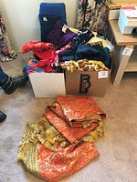 Two boxes and a folded reddish-gold bedspread with gold tassels on floor among fabric remnants
