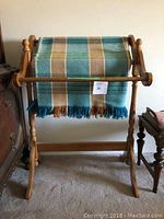 Wooden blanket and quilt stand with a multi-colored woven blanket draped over the middle horizontal bar, positioned between two pieces of wooden furniture.