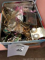Overhead view of open metal tin filled with assorted vintage costume jewelry including earrings, necklaces, bracelets, brooches packed loosely and in plastic bags.