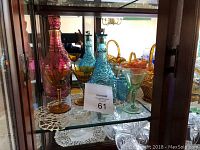 Display shelf showing coloured glass decanters and glasses of pink, blue, amber, and green hues along with an inventory card.