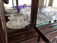 Wide view of bottom shelf display cabinet showing multiple crystal pitchers, bowls, and glassware arranged on lace doilies.