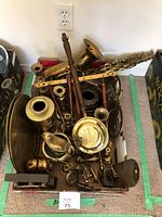 Photo showing a box full of brass items including part of a saxophone body with wooden handles, candlesticks, brass trays, and various decorative figurines and objects.