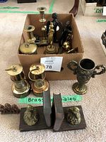 Photo showing assortment of brass collectibles including candle holders, lanterns, bookends with figurines, and trophy cup arranged on carpet and in cardboard box.