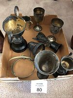 Photo showing ten various antique trophies and cups in a cardboard box, displaying metal trophies with patina and a large gold-toned interior trophy.