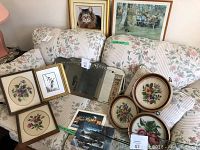 Photo showing multiple framed floral needlepoint pieces, a framed cat print, a vintage mirror, and decorative floral plates on a floral upholstered couch.