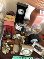 Overview of clocks of various styles and conditions including a tall black pendulum clock, red clock, brass clock, and small decorative clocks arranged on table near lamp.