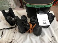 Photo showing the three binoculars on a surface with a black carrying case labeled 'Binoculars'. Three binoculars include Kurt Muller, S Riefler, and a smaller binocular labeled Made in China 3x30.