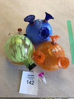 Three handblown glass hummingbird feeders arranged on a beige surface showing green, blue, and orange feeders with multiple feeding ports and color accents.