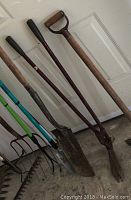 View of long handled shovel, spade, and Wilkinson Sword edging shears with wooden handles leaning against white door.