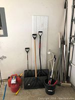 View of snow shovels, gas cans, bucket with squeegee, pole tools, and car top rails leaning against the wall.