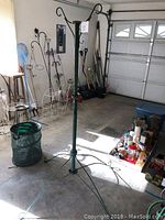 Full view of the tall green metal hanging basket holder standing in a garage with a tripod base and three hooks at the top.