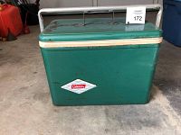 Front view of green Coleman metal cooler with diamond logo, white trim, and attached metal handle folded down, showing rust and wear.