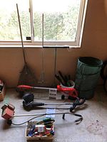 View of two electric trimmers (Shopcraft and B&D) on the floor under window, various garden hand tools and box of smaller tools beneath.