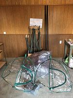 Greenhouse parts including green tubular metal frames and clear plastic sheeting with two long black poles leaning against the wall