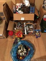 Overview of boxes and items containing Christmas decorations such as figurines, floral arrangements, and glassware on wooden floor.