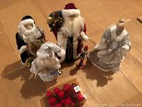 Four standing Santa figurines in various festive outfits with a white and silver angel figurine and a box of red ball ornaments on a wooden floor.