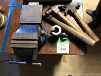 Photo showing a bench vise, two claw hammers with wooden handles, and two metal cylindrical components with clamps placed on a wooden table.