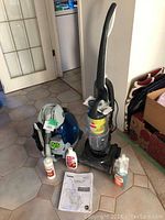 Two vacuum cleaners—Hoover Steam Vac Duo carpet cleaner and Bissell Prolite Compact upright vacuum—placed on tiled floor with cleaning solution bottles and user manual.