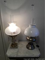 Photo of two electric hurricane lamps on a marble surface against a white wall, showing details of the glass shades, metal bases, and electric cords.
