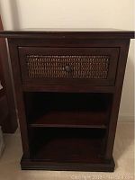 Front view of a single side table showing drawer with woven front panel, open shelf space below, and dark wood finish.