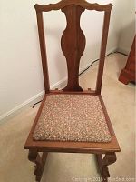 Wooden rocking chair with floral cushioned seat on a carpeted floor against a white wall.